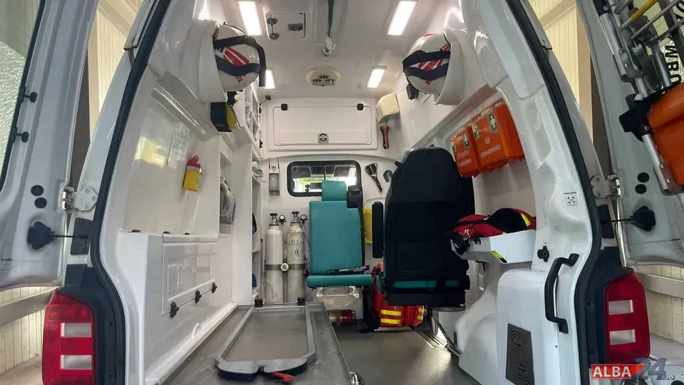 Interiorul unei ambulanțe cu uși deschise, echipată cu o butelie de oxigen, un scaun de culoare turcoaz și un pat de transport metalic.