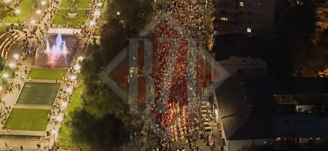 O mulțime de oameni merge pe un drum îngust între copaci și clădiri, lângă un parc cu fântâni luminoase sub un cer de noapte.