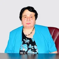 O femeie cu părul scurt și negru poartă un sacou albastru peste o bluză neagră cu motive florale și un colier de perle negre.