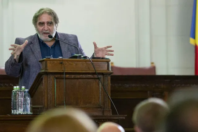 Un bărbat cu barbă și părul cărunt stă în spatele unui podium de lemn, având mâinile ridicate și gura deschisă.