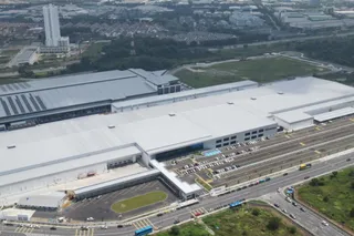 O vedere aeriană prezintă un complex industrial de mari dimensiuni cu acoperișuri plate și gri, situat lângă o autostradă și o zonă de parcare.