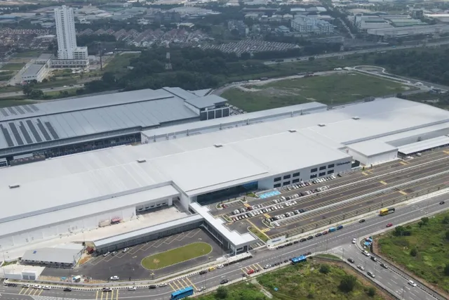 O vedere aeriană prezintă un complex industrial de mari dimensiuni cu acoperișuri plate și gri, situat lângă o autostradă și o zonă de parcare.