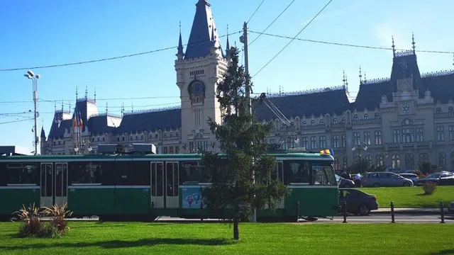 Un tramvai verde și alb circulează pe o stradă lângă un copac și un teren cu iarbă verde, având o clădire mare în fundal.