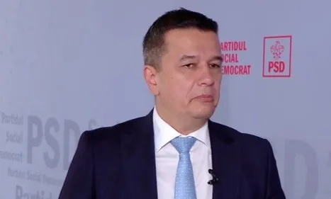 Un bărbat cu părul scurt, purtând sacou închis la culoare, cămașă albă și cravată albastră, stă în fața unui fundal cu inscripții și un logo roșu.