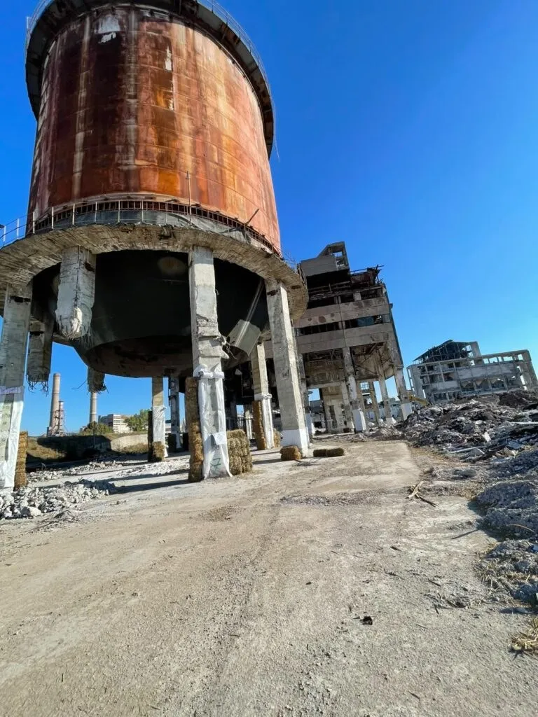 Un rezervor cilindric ruginit sprijinit pe stâlpi, în fața unei construcții dezafectate și a unor ruine sub cer senin.
