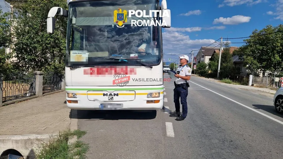 Un polițist în uniformă stă pe marginea unui drum lângă un autocar alb cu detalii verzi și un logo colorat.