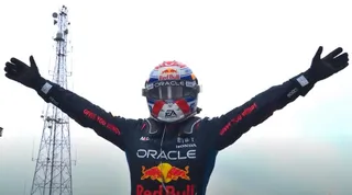 Un pilot de curse cu mâinile ridicate poartă un costum de protecție negru și un cască colorat cu logourile Red Bull și Oracle.