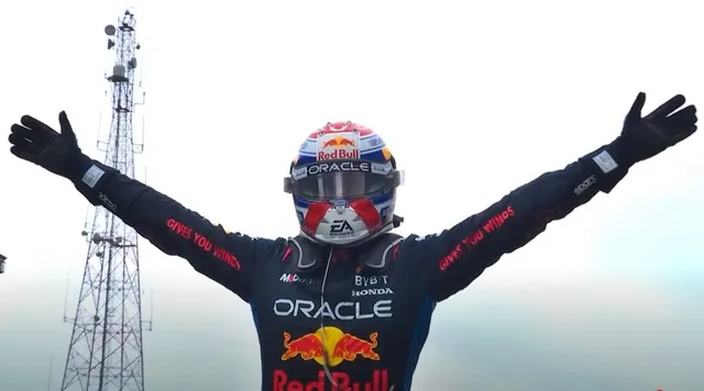 Un pilot de curse cu mâinile ridicate poartă un costum de protecție negru și un cască colorat cu logourile Red Bull și Oracle.