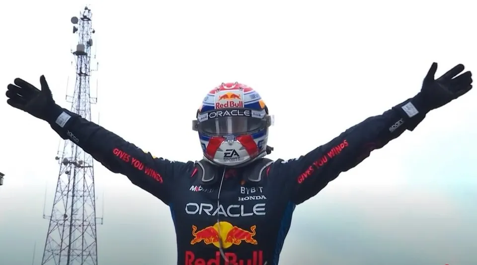 Un pilot de curse cu mâinile ridicate poartă un costum de protecție negru și un cască colorat cu logourile Red Bull și Oracle.
