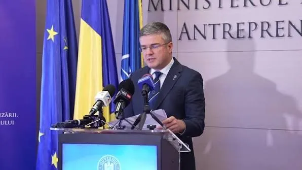 Un bărbat cu ochelari și costum închis la culoare stă în spatele unui podium cu microfoane, în fața unor steaguri.
