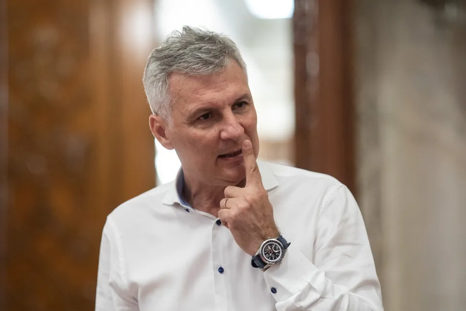 Un bărbat cu părul scurt și cărunt poartă o cămașă albă și își ține un deget pe obraz sub ochelari, purtând un ceas la încheietura mâinii.