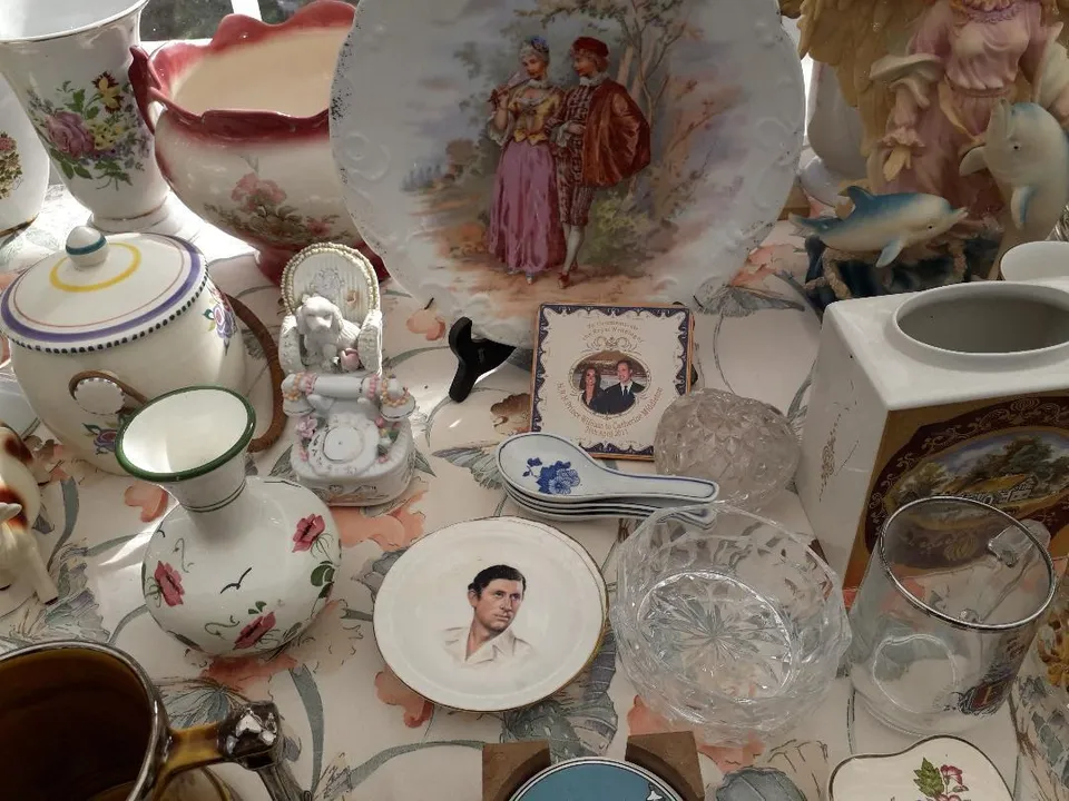 Diverse obiecte de ceramică decorativă, inclusiv farfurii cu figuri umane, vaze florale și boluri de cristal, sunt dispuse pe o suprafață acoperită cu o față de masă.