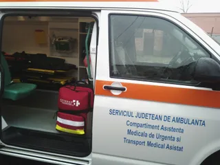 Interiorul unei ambulanțe cu echipamente medicale și o geantă roșie de transport aflată lângă scaunul verde.