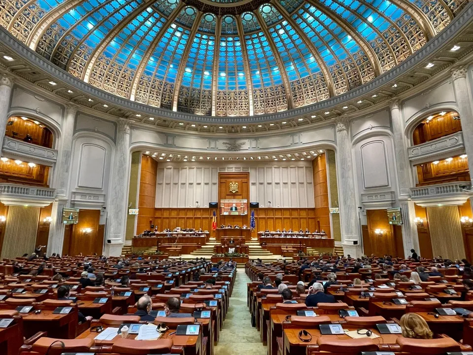 Imaginea prezintă o sală de plen a unei instituții legislative, cu numeroase bănci din lemn ocupate parțial de persoane, sub o cupolă vitrată.