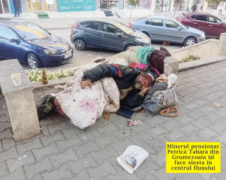 Un bărbat stă întins pe un trotuar lângă un beton, acoperit cu o pătură cu modele florale și purtând haine colorate.