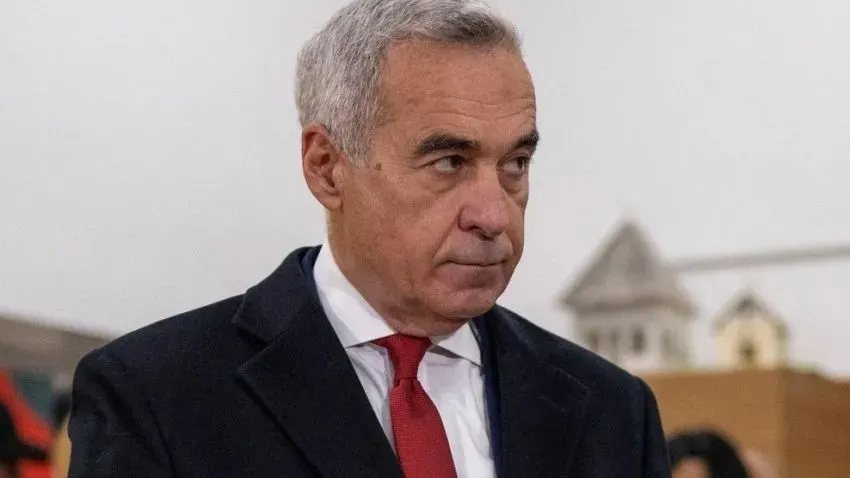 Un bărbat cu părul cărunt, purtând sacou închis la culoare, cămașă albă și cravată roșie, privește spre lateral într-un decor cu fundal deschis.
