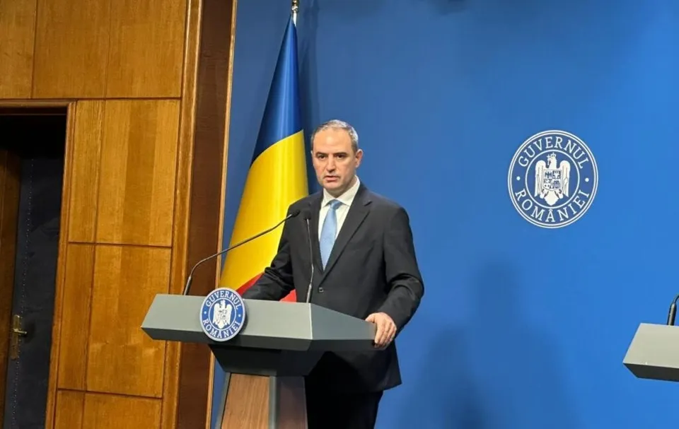 Un bărbat într-un costum închis la culoare și cravată deschisă stă în spatele unui podium gri în fața unui steag și a unui fundal albastru.
