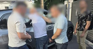 Patru bărbați stau lângă o mașină gri, unul purtând un tricou alb și pantaloni albaștri, iar altul un tricou negru cu modele florale.