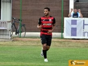 Un bărbat cu tricou roșu și negru, pantaloni scurți negri și șosete negre aleargă pe un teren de fotbal cu iarbă verde.
