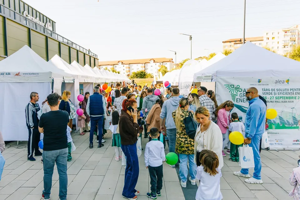 O mulțime de oameni stă în picioare pe o esplanadă pavată între mai multe corturi albe de expoziție sub un cer luminos.