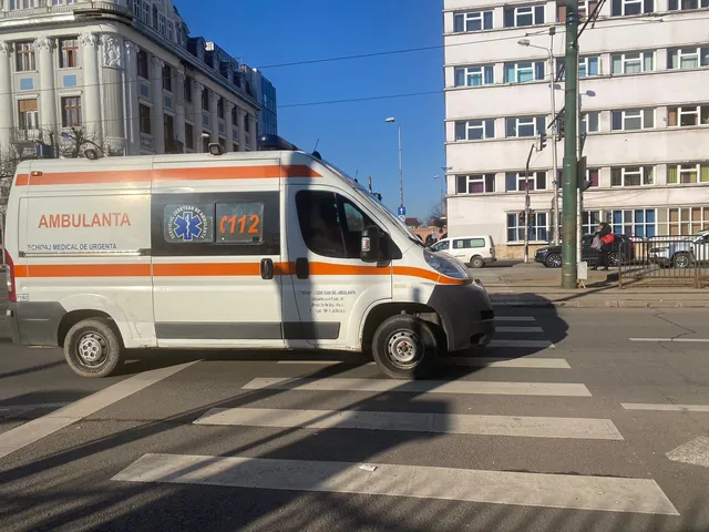 O ambulanță albă cu dungi portocalii este oprită pe un drum asfaltat lângă un zebru, în fața unor clădiri urbane sub un cer senin.