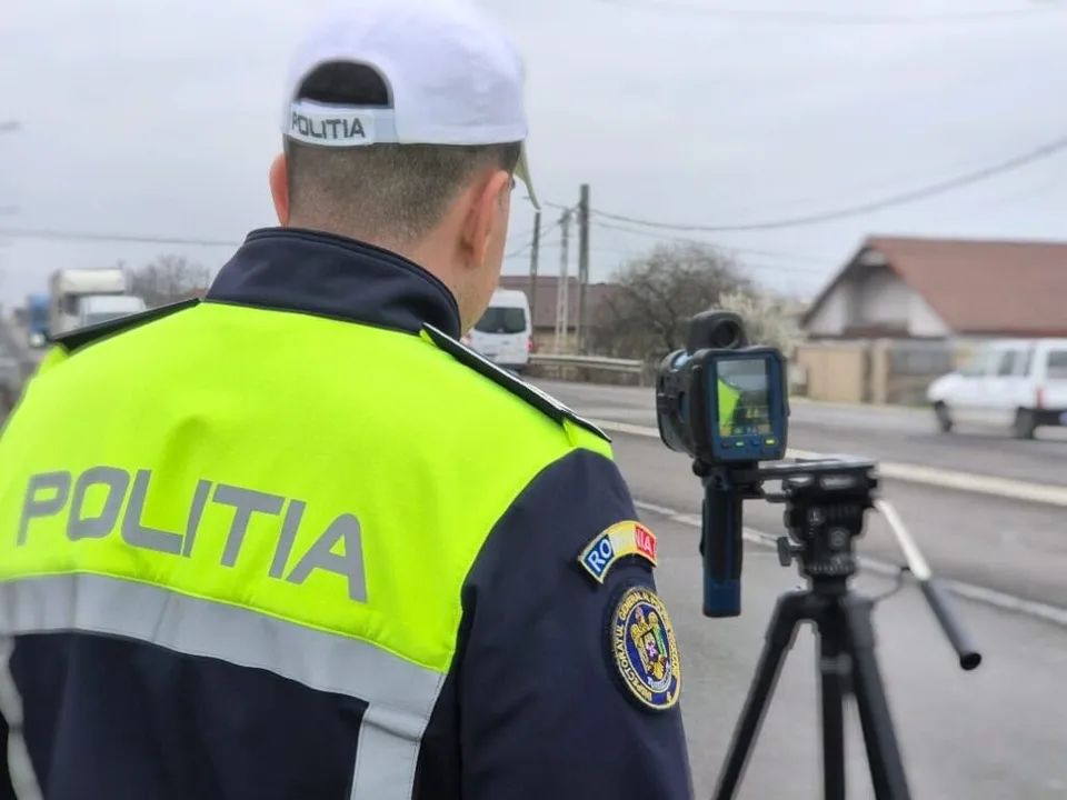 Un agent de poliție cu uniformă reflectorizantă stă lângă o cameră video montată pe un trepied pe marginea unui drum.