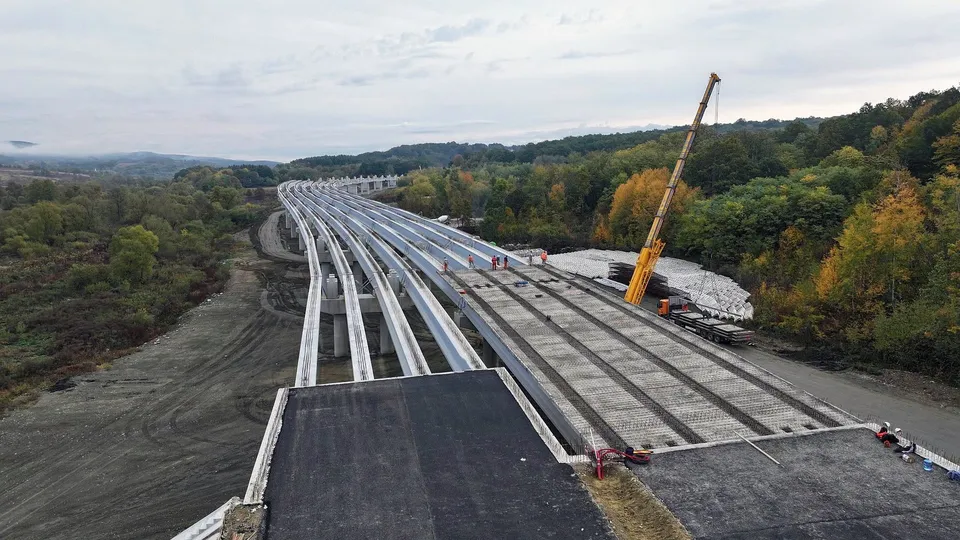 O vedere de sus a unei structuri de viaduct în construcție cu piloni albi și suprafețe de beton, lângă o zonă cu vegetație și un excavator.
