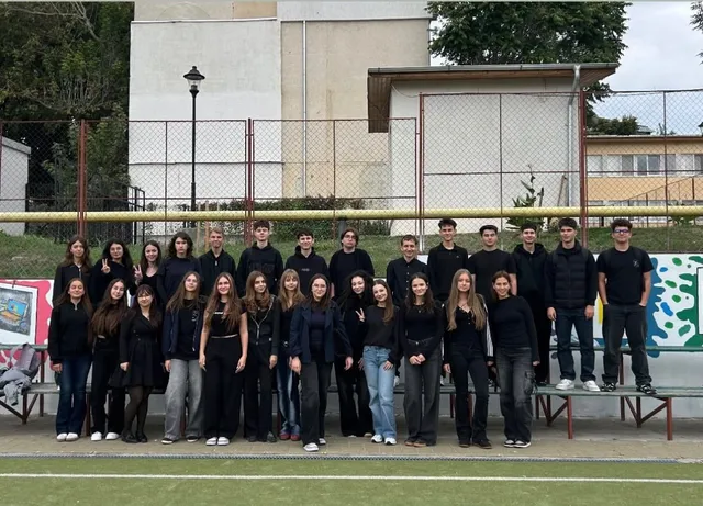 Un grup de tineri îmbrăcați în haine negre stă poziționat pe o bancă de lemn în fața unui gard metalic și al unei clădiri albe.