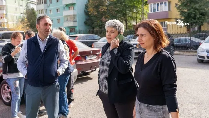 Un grup de persoane stă pe o stradă lângă mașini parcate, în timp ce o femeie vorbește la telefon mobil.