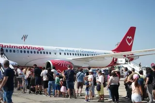 O mulțime de oameni stă în picioare pe o platformă de aeroport lângă un avion alb cu inscripții roșii și un coadă roșie.