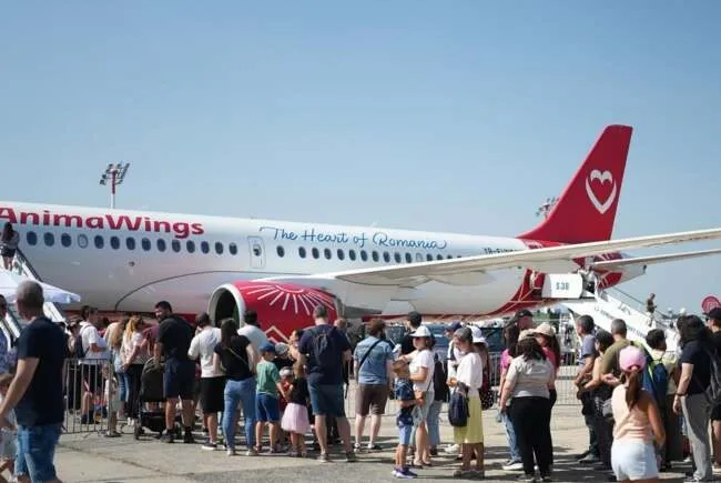 O mulțime de oameni stă în picioare pe o platformă de aeroport lângă un avion alb cu inscripții roșii și un coadă roșie.