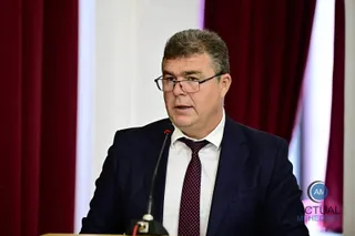 Un bărbat cu ochelari, purtând sacou închis la culoare, cămașă albă și cravată bordeaux, stă în fața unui microfon.