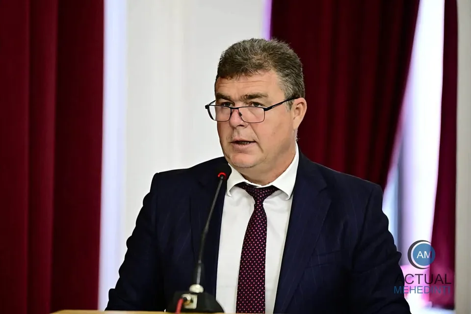 Un bărbat cu ochelari, purtând sacou închis la culoare, cămașă albă și cravată bordeaux, stă în fața unui microfon.