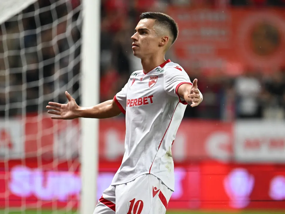 Un fotbalist într-un echipament alb cu detalii roșii stă cu brațele întinse pe un teren de fotbal, lângă o plasă de porți.
