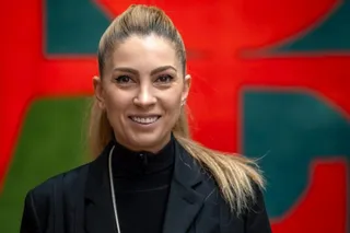 O femeie cu părul blond strâns într-o coadă de cal poartă un sacou negru și un guler negru în fața unui fundal colorat.