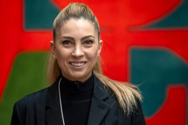 O femeie cu părul blond strâns într-o coadă de cal poartă un sacou negru și un guler negru în fața unui fundal colorat.