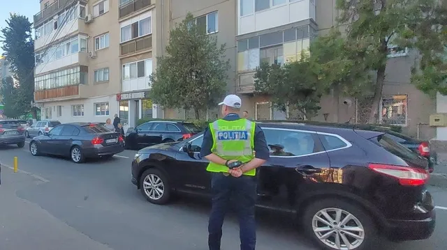 Un polițist cu vestă reflectorizantă verde stă cu spatele lângă o mașină neagră parcată pe un drum urban lângă clădiri de apartamente.