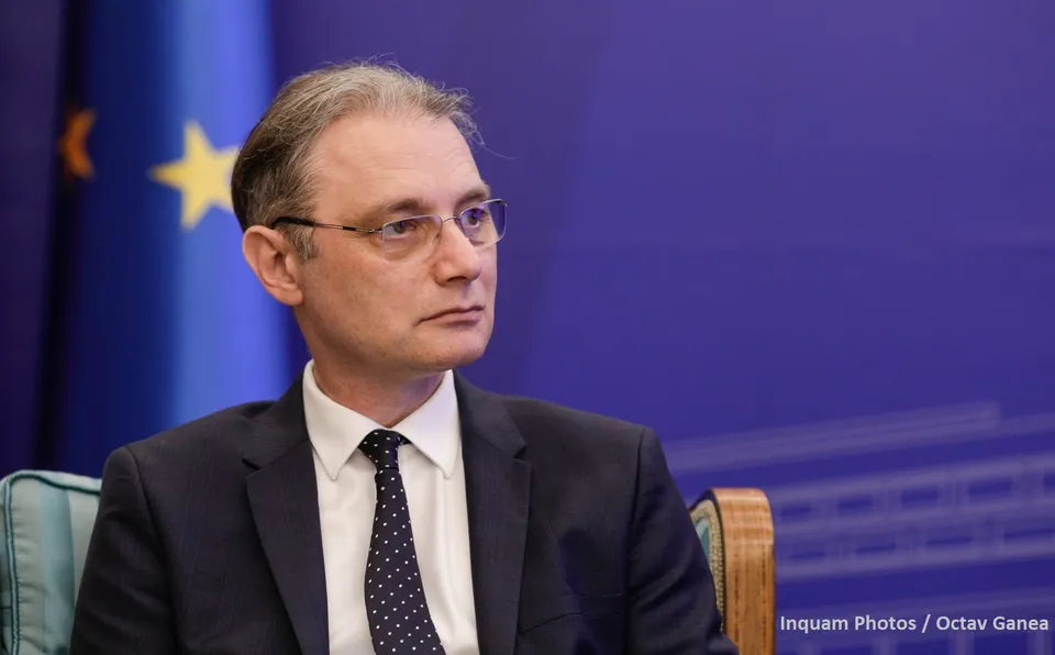 Un bărbat cu ochelari, purtând sacou închis și cravată cu puncte albe, stă în fața unui fundal albastru cu o stea galbenă.