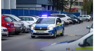 O mașină de poliție albă cu lumini albastre aprinse circulă pe un drum asfaltat printre alte vehicule parcate.