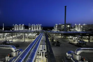 O vedere de sus asupra unei instalații industriale cu conducte metalice strălucitoare, turbine și un coș de fum înalt sub un cer albastru înserat.