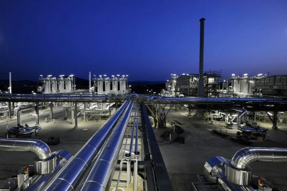 O vedere de sus asupra unei instalații industriale cu conducte metalice strălucitoare, turbine și un coș de fum înalt sub un cer albastru înserat.
