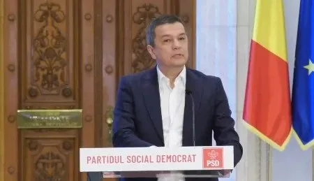 Un bărbat cu cămașă albă și sacou închis stă în spatele unui podium alb cu inscripția PARTIDUL SOCIAL DEMOCRAT.