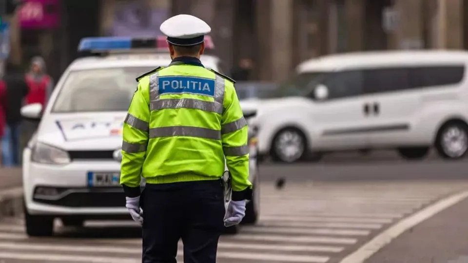 Un polițist cu vestă reflectorizantă scrisă POLITIA stă cu spatele pe o stradă lângă un vehicul alb și un zebru.
