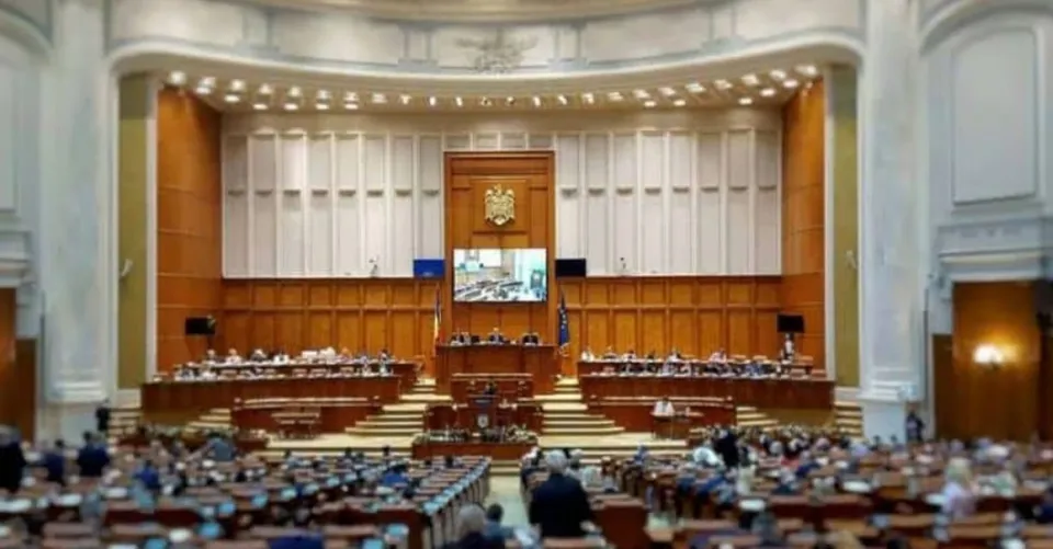 Sală parlamentară cu tribune, pupitre, ecrane, steme și numeroși oameni așezați în plan frontal și central.