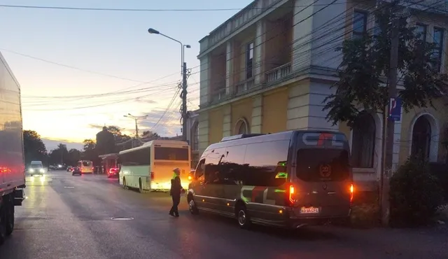Imaginea prezintă o stradă aglomerată la apus, cu un polițist local în prim-plan, inspectând un microbuz. Pe stradă se află mai multe autobuze și camioane.