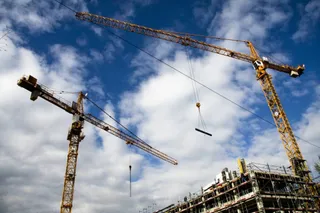 Două macarale de construcții galbene se ridică pe un cer albastru cu nori plutind deasupra unei structuri de clădire în lucru.