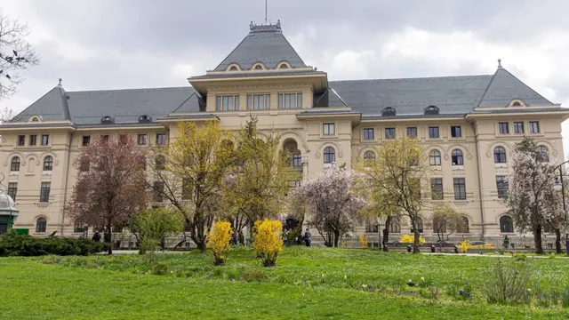 O clădire mare de culoare deschisă cu acoperiș gri înconjurată de copaci cu frunze verzi și roz pe un teren cu iarbă verde.
