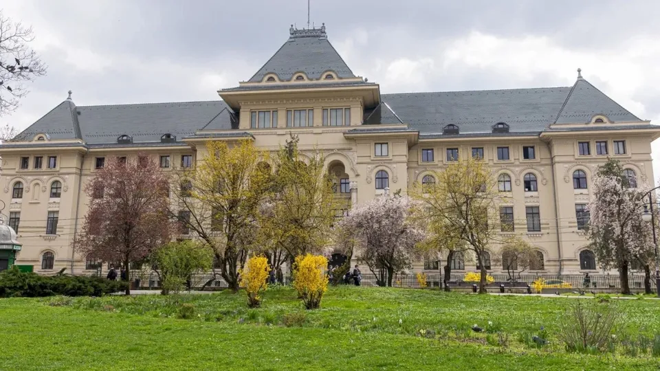 O clădire mare de culoare deschisă cu acoperiș gri înconjurată de copaci cu frunze verzi și roz pe un teren cu iarbă verde.