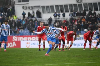 Câțiva jucători de fotbal în echipamente albastre și roșii dispută un meci pe un teren de gazon verde sub un cer înnorat.