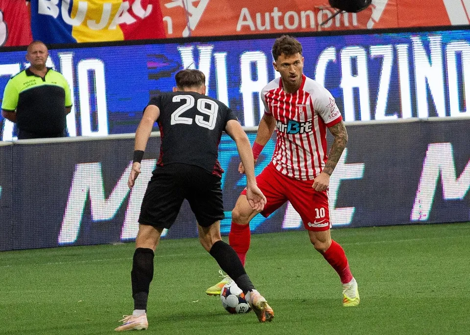 Un jucător în echipament roșu și alb conduce mingea pe terenul de fotbal în fața unui adversar care poartă tricou negru cu numărul 29.